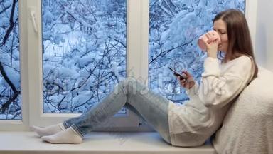 漂亮的女孩坐在窗台上，<strong>喝茶</strong>，用智能手机。 <strong>冬天</strong>在外面。